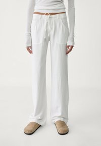 Białe szerokie spodnie z elastycznym pasem ze sznurkiem, dwoma przednimi kieszeniami i miękką fakturą materiału, zestawione z beżowymi wsuwanymi butami.