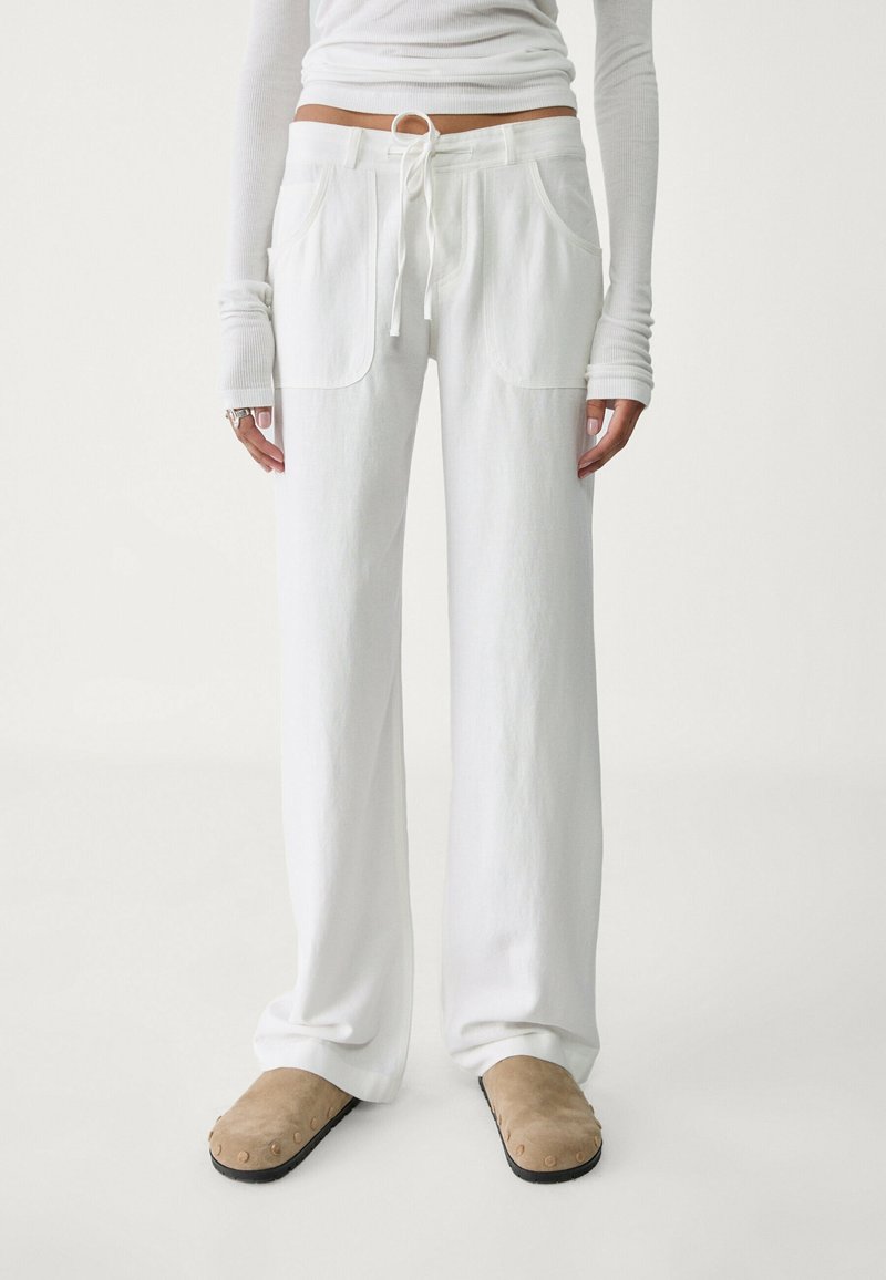 Białe szerokie spodnie z elastycznym pasem ze sznurkiem, dwoma przednimi kieszeniami i miękką fakturą materiału, zestawione z beżowymi wsuwanymi butami.