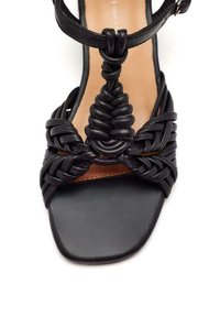 Sandalia de cuero negra con un intrincado diseño trenzado, puntera cuadrada y un destacado acento trenzado en el centro. Incluye una correa ajustable.