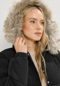 Chaqueta acolchada negra con capucha de piel sintética. Textura suave con cierre de cremallera y herrajes plateados. Cálida e insulated para el clima frío.