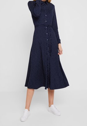 Woman wearing a navy blue long-sleeve midi dress with white polka dots, front buttons, and white sneakers, standing against a plain backdrop.