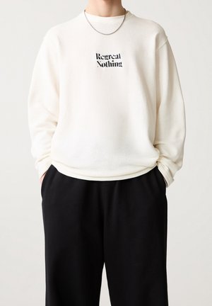 Personne portant une chemise blanche à manches longues avec le texte « Regret Nothing » et un pantalon noir, les mains dans les poches, devant un fond clair uni.