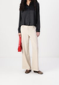 Donna che indossa una blusa nera a maniche lunghe, pantaloni beige a gamba larga, scarpe ballerine con stampa leopardata, mentre tiene una piccola borsa rossa su uno sfondo bianco semplice.