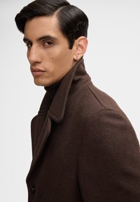 Bruine wollen jas met een hoge kraag, dubbelzijdige knopen en een gestructureerde stof. Heeft een moderne snit. Model draagt eronder een donkere turtleneck.