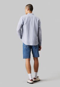 Camicia azzurra a righe con bottoni e maniche lunghe, abbinata a pantaloncini di jeans blu. Il modello indossa calzini bianchi e sneakers in tonalità neutra.