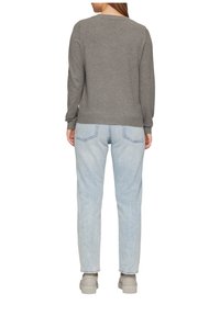 Kvinna iklädd grå långärmad tröja, ljusblå jeans och beiga sneakers, står vänd med ryggen mot en vit bakgrund.