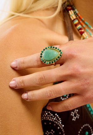 Bague en or ornée d'une grande pierre verte ovale entourée de petites pierres ressemblant à des émeraudes. Montre une main texturée avec des ongles rose clair.