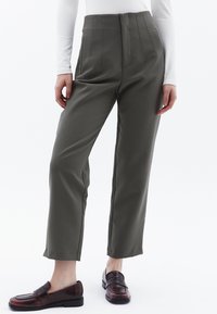 Olivgrön tapered byxor med hög midja, veck och sidofickor, kombinerat med mörkbruna slip-in skor med en strukturerad yta.