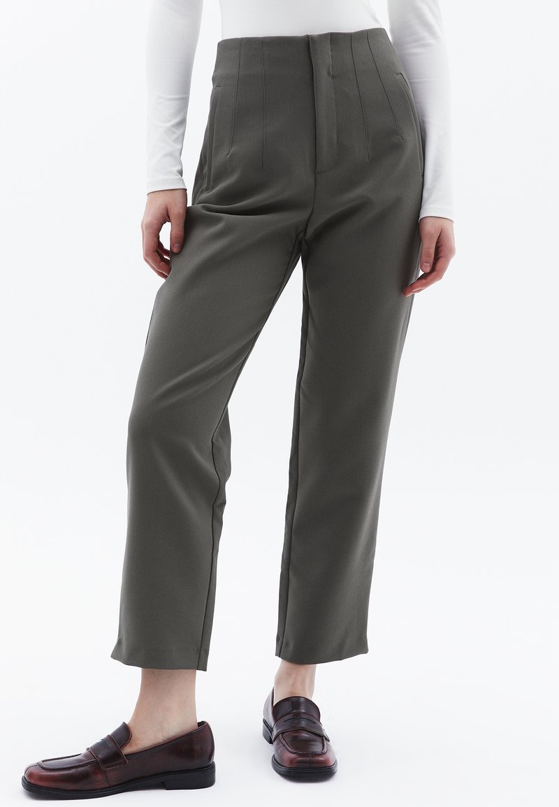 Oliven grøn tapered bukser med høj talje, læg og sidelommer, parret med mørkebrun slip-on sko, der har en tekstureret finish.