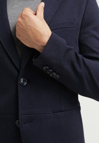 Navy blazer with a textured fabric, featuring a single breast pocket and four-button cuffs. Includes round buttons and a tailored fit.