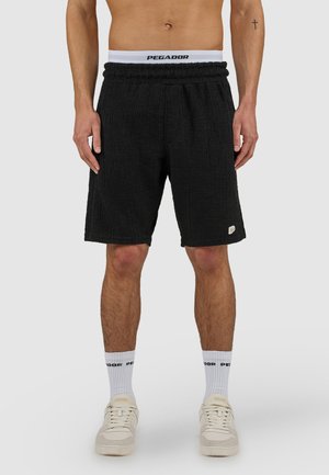 Hombre con pantalones cortos negros texturizados, calcetines blancos con el texto "PEGADOR" y zapatillas blancas, de pie contra un fondo gris liso.