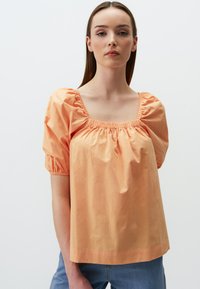Blouse en coton couleur pêche avec un décolleté carr�é, des détails froncés et des manches bouffantes. Portée avec un jean en denim clair. Texture lisse.