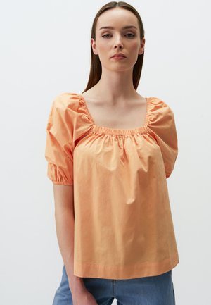Blouse en coton couleur pêche avec un décolleté carré, des détails froncés et des manches bouffantes. Portée avec un jean en denim clair. Texture lisse.