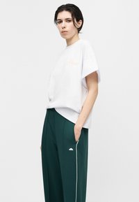 Άτομο με κοντά σκούρα μαλλιά, που φοράει λευκό υπερμεγέθη μπλουζάκι και σκούρα πράσινα παντελόνια φαρδιά με λευκή επένδυση και λογότυπο.