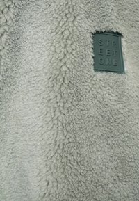 Fleece-material i ljusgrått, med en texturerad, fluffig yta. Inkluderar en liten, fyrkantig, grön etikett präglad med "STREET ONE."
