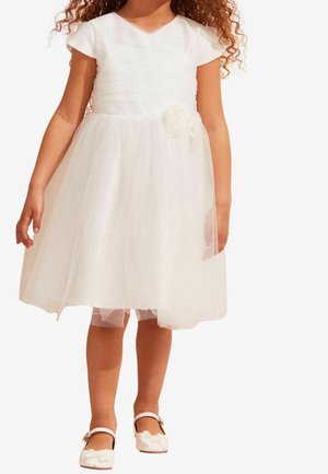 Weißes Blumenkleid mit tailliertem Oberteil und Tüllrock. Kurzärmel und ein Blumenakzent an der Taille. Kombiniert mit weißen Schuhen.