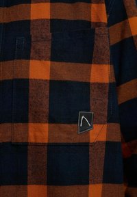 Flanellen overhemd met een marineblauw en oranje geruit patroon, voorzak met een zwarte vierkante patch en wit logo, textuur van katoen.
