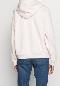 Světle růžová mikina s velkou kapucí, sklopenými rameny a žebrovaným lemem. Nosená s modrými džíny, které zdůrazňují uvolněný střih a jemnou texturu.
