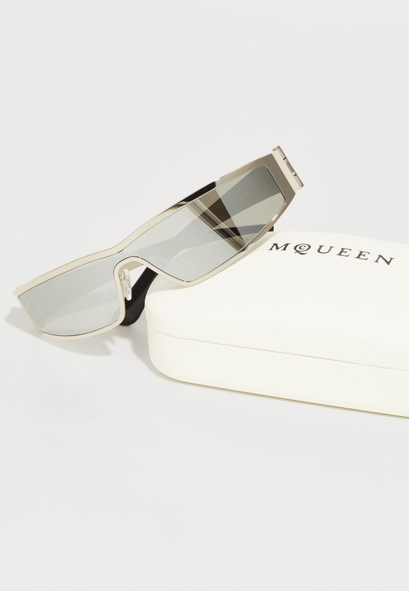 Silberne Sonnenbrille mit rechteckigen, grauen Gläsern. Enthält ein weißes Etui mit der Aufschrift "MQUEEN" in schwarzer Schrift.