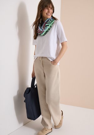 Nuori nainen hymyilee, pukeutuneena valkoiseen lyhythihaiseen paitaan, beigenvärisiin leveälahkeisiin housuihin, moniväriseen huiviin, pitää mustaa kangaskassia, seisoo vaaleaa seinää vasten.