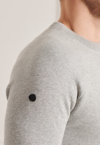 Maglione grigio in maglia con collo rotondo e una toppa nera con logo sulla spalla. La texture sembra morbida e liscia.