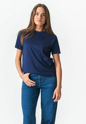 Kvinde med langt brunt hår iført en marineblå t-shirt og blå jeans, stående mod en ensartet hvid baggrund.