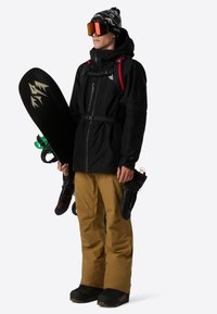 Sort vandtæt jakke med røde detaljer, lysebrune snebukser, sorte snowboardstøvler. Holder en sort snowboard, sorte vanter og en rygsæk.