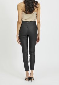 Femme avec de longs cheveux noirs portant un haut sans manches beige, un pantalon noir ajusté taille haute avec fermeture éclair au dos, et des mules à talons noires, debout.