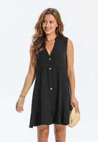 Robe noire sans manches avec un col en v, fermeture boutonnée sur le devant, ourlet à volants superposés et tissu texturé. Accessoirisée avec un petit sac tissé.