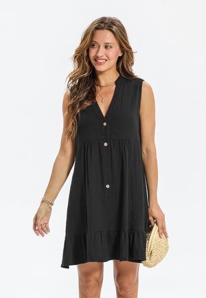 Robe noire sans manches avec un col en v, fermeture boutonnée sur le devant, ourlet à volants superposés et tissu texturé. Accessoirisée avec un petit sac tissé.