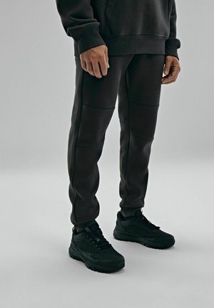 Czarny zestaw odzieży sportowej składający się z bluzy typu crew neck oraz tapered joggerów z detalami w postaci paneli, zestawiony z czarnymi butami sportowymi.