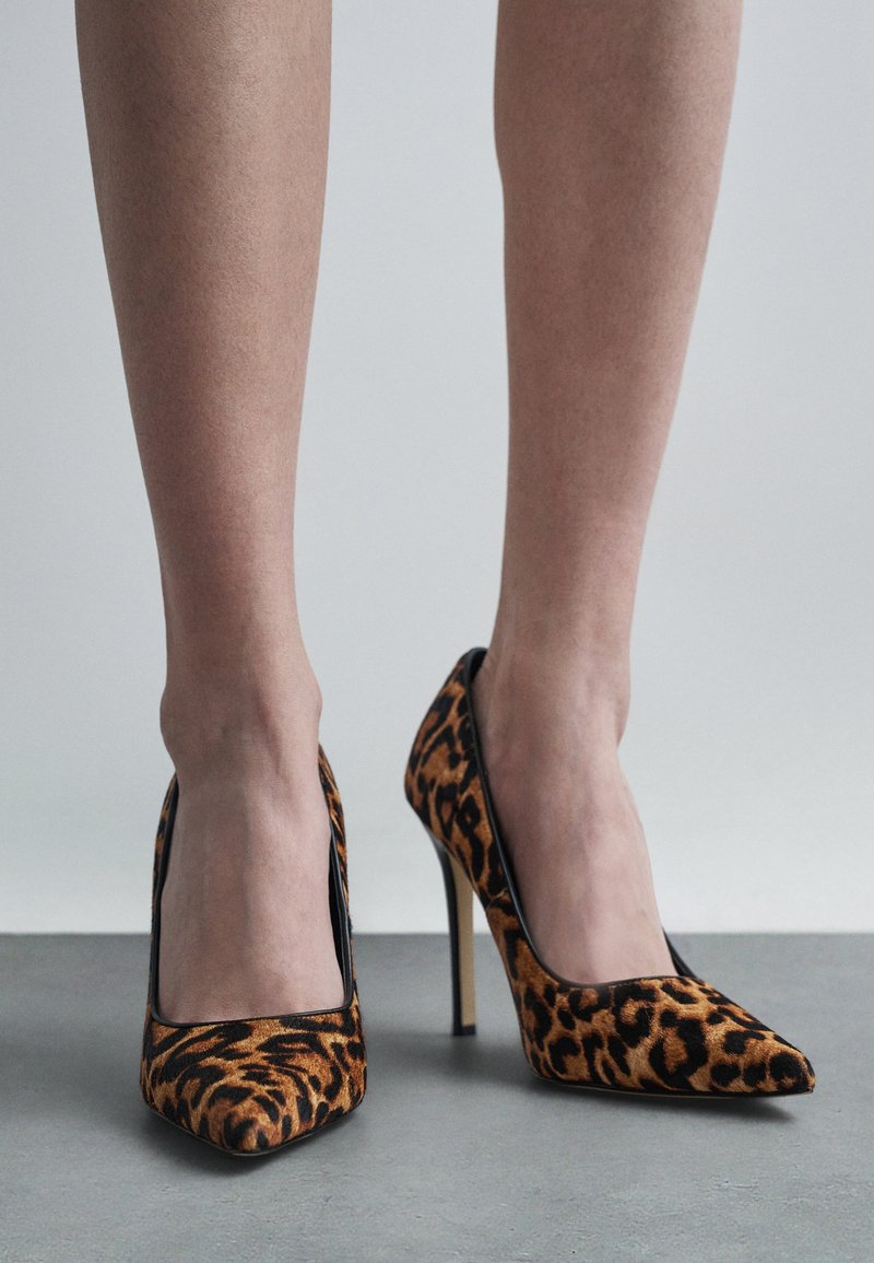 Noge v togem čevlju z ostrim nosom in leopardjim vzorcem, ki stojijo na gladkem sivi tlaku proti enotnemu svetlemu ozadju.