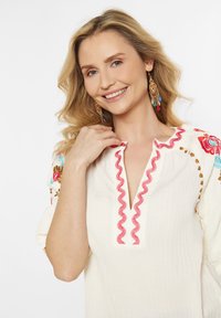 Top alb texturat cu guler roșu în formă de valuri, având broderie florală în nuanțe de albastru, roz și maro pe mâneci. Cercei colorati cu pandantive.