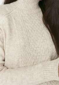 Hellbeiger gestrickter Pullover mit hohem, geripptem Kragen und Zopfmuster auf der Vorderseite. Verfügt über lange Ärmel und eine strukturierte Stoffoberfläche.