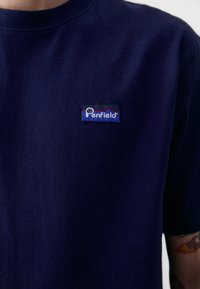 Tricou din bumbac bleu marin, cu guler rotund și mâneci scurte, având un patch cu logo-ul Penfield cusut pe piept.