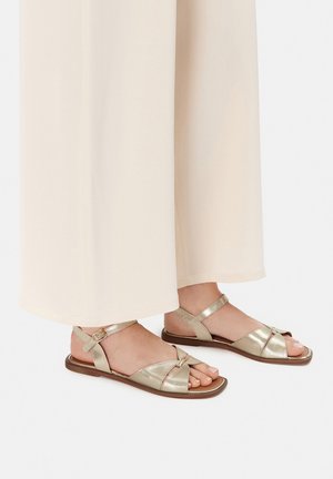 Złote metaliczne sandały o fakturowanym wykończeniu, z odkrytymi palcami, regulowanym paskiem wokół kostki i płaską podeszwą, zestawione z jasnymi spodniami.