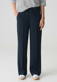 Marineblauwe plooibroek van soepele stof, met een trekkoord in de taille, zijzakken, en gecombineerd met witte sneakers en een gestreept shirt.