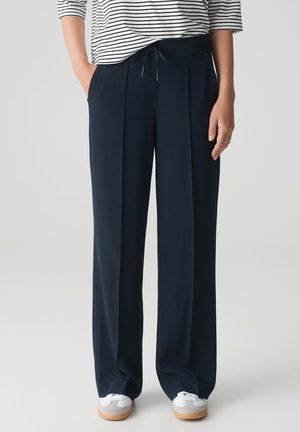 Navy pleated trousers made of smooth fabric, featuring a drawstring waist, side pockets, and paired with white sneakers and a striped top.