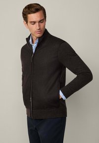 Veste zippée marron foncé en tissu texturé, avec un col haut et des poches latérales ; portée par-dessus une chemise bleu clair avec un col visible.