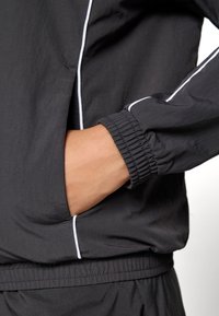 Mão dentro do bolso de uma jaqueta preta com detalhes brancos, punhos elásticos e calças pretas combinando com uma cintura elástica.