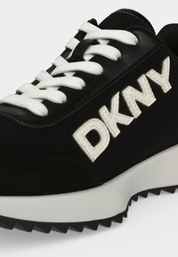 Baskets noires et blanches avec lacets blancs et grand logo DKNY en relief sur le côté, avec une semelle extérieure noire à rainures et une semelle intermédiaire blanche.