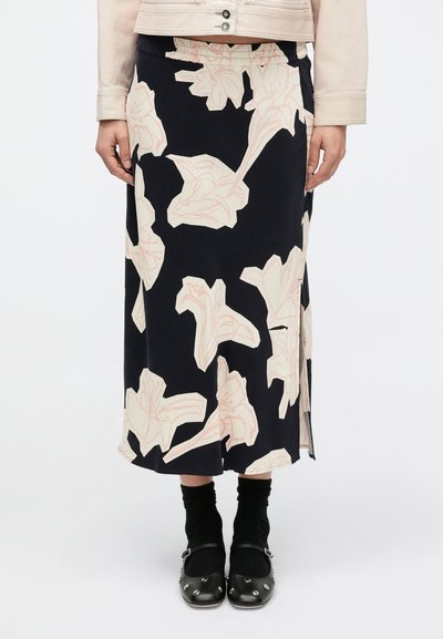 Jupe midi à motif floral en bleu marine foncé avec de grandes fleurs roses, confectionnée dans un tissu doux. Dispose d'une fente latérale pour faciliter les mouvements. Portée avec des chaussettes et des chaussures noires.
