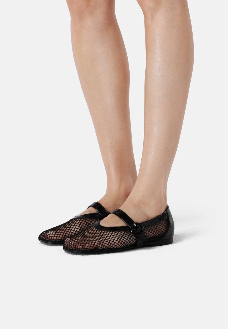 Jambes portant des ballerines Mary Jane en résille noire avec une bride à boucle ronde sur le pied, sur fond blanc uni.