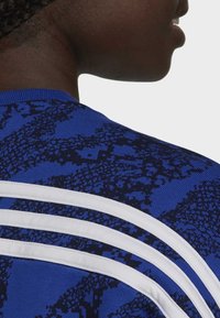 Camisa atlética azul con un patrón texturizado, cuello redondo y tres franjas blancas en la zona de los hombros en la parte posterior.
