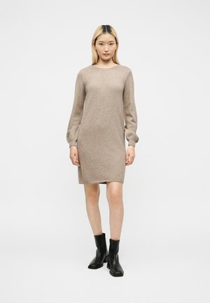 Jonge vrouw met steil blond haar die een beige gebreide jurk met lange mouwen en zwarte enkellaarzen draagt, staand tegen een effen witte achtergrond.