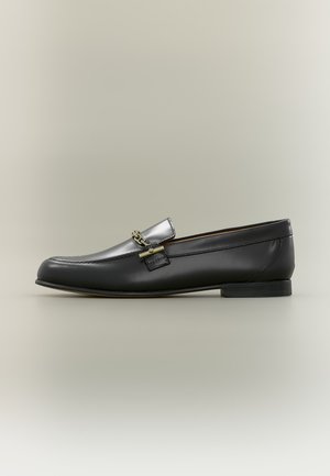Zwarte leren loafer met zilveren bovenkant, gouden kettingdetail, lage hak en afgeronde neus op een neutrale achtergrond.