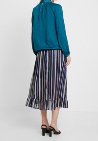 Blouse teal à manches longues, décolleté froncé, associée à une jupe midi rayée navy et blanche avec un ourlet volanté et des talons hauts.