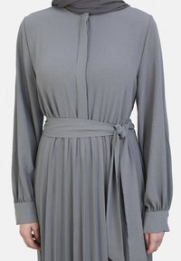 Graues Langarmkleid mit plissiertem Rock, Stoffbindegürtel in der Taille, weicher Textur und Knöpfen am Ausschnitt. Akzentuiert mit einem passenden grauen Schal.