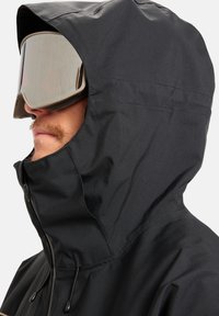 Juodas slidinėjimo džemperis su aukštu apykakle ir reguliuojama kapišonu, pasižymintis lygiu tekstūru. Pridėti dideli veidrodiniai slidinėjimo akiniai.