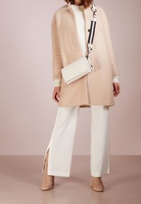 Cappotto oversize beige in tessuto testurizzato, maglione bianco sotto, pantaloni crema con fessure laterali e stivali beige alla caviglia. Borsa a tracolla bianca.